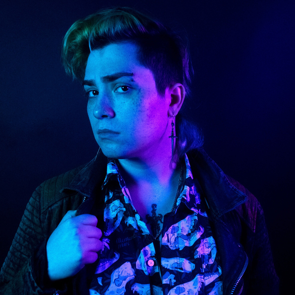 Joan wearing a skull buttoned shirt, with their left hand up to their jacket looking to camera in bisexual lighting.